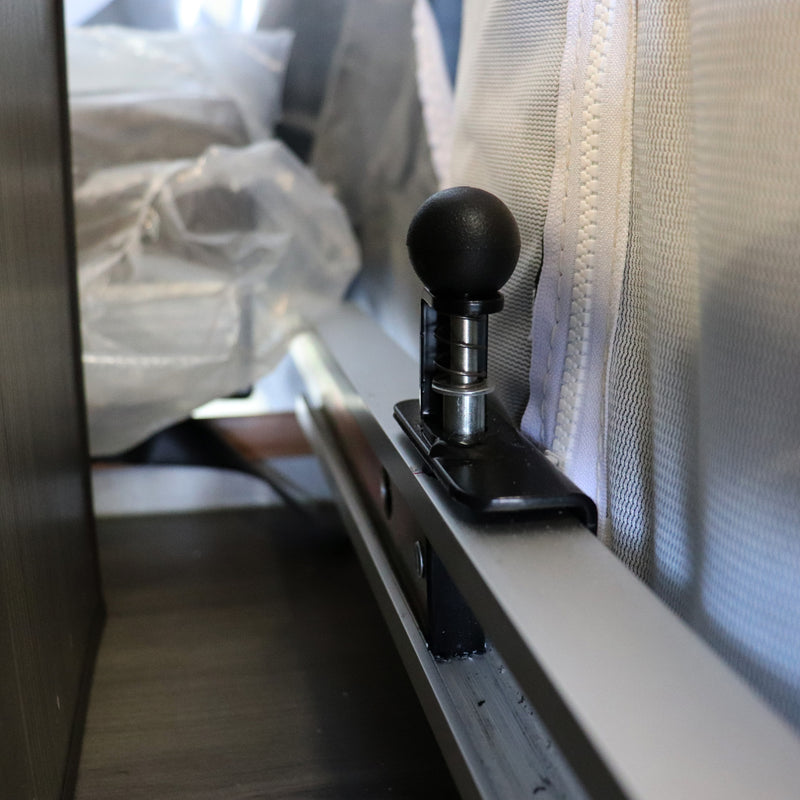 A black metal bed rail plunger mounted on a white surface, part of a camping trailer's bed locking mechanism.
