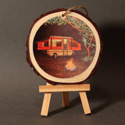 A circular wooden ornament displaying a chalk art print of a red camper trailer with trees and a campfire in the background, mounted on a small wooden stand.