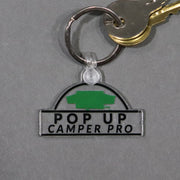 A clear acrylic keychain with a green and black print reading 'POP UP CAMPER PRO'. It is attached to a ring with a set of keys.