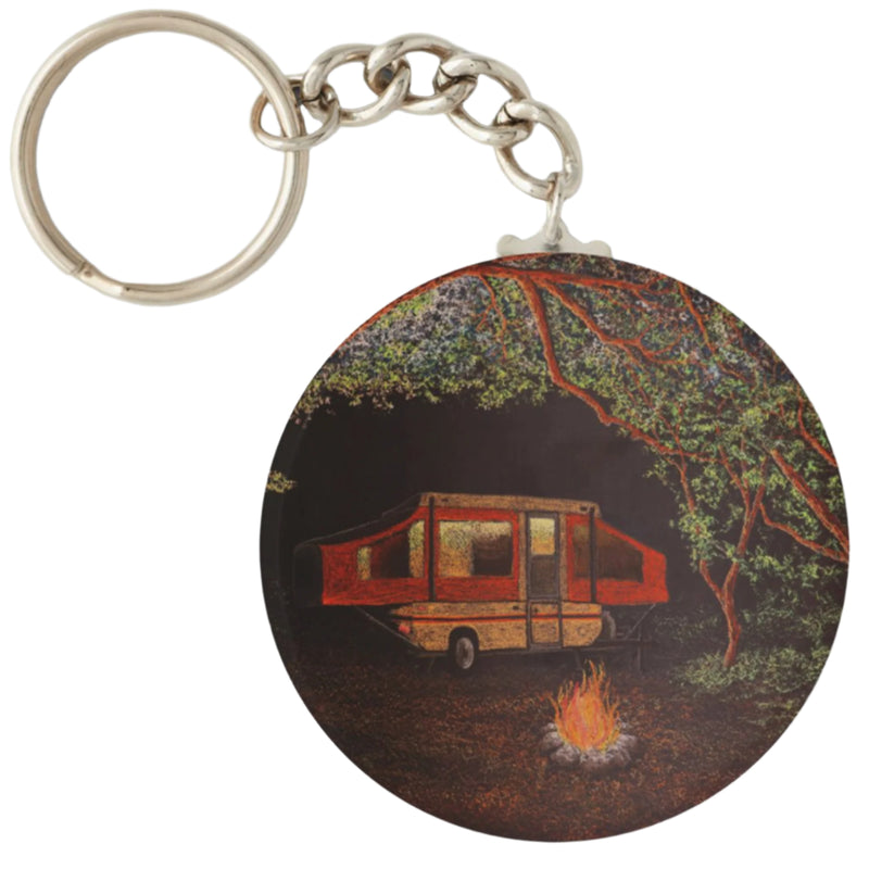 A round keychain with a printed artwork of a camping scene, featuring an RV and a campfire, with a tree in the background.