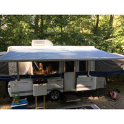 A white patio awning installed on a camper, providing shade over the outdoor area. The awning is extended out from the side of the camper, which is parked in a campsite with trees in the background.